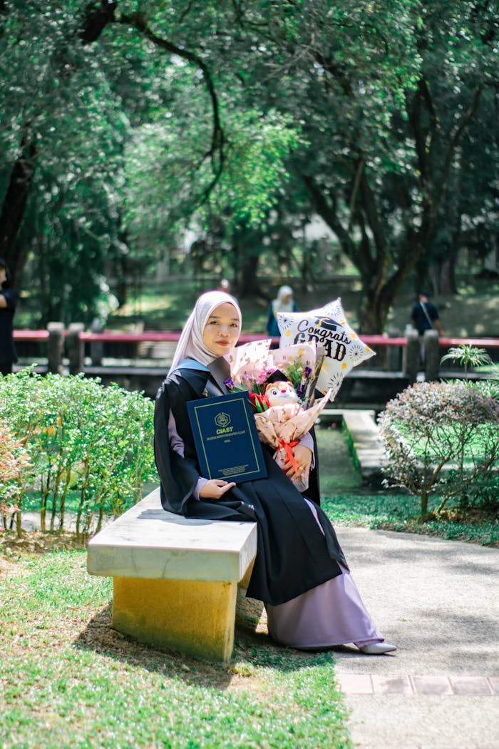 A woman in graduation attire sitting on a bench with balloons and diploma in Shah Alam park.