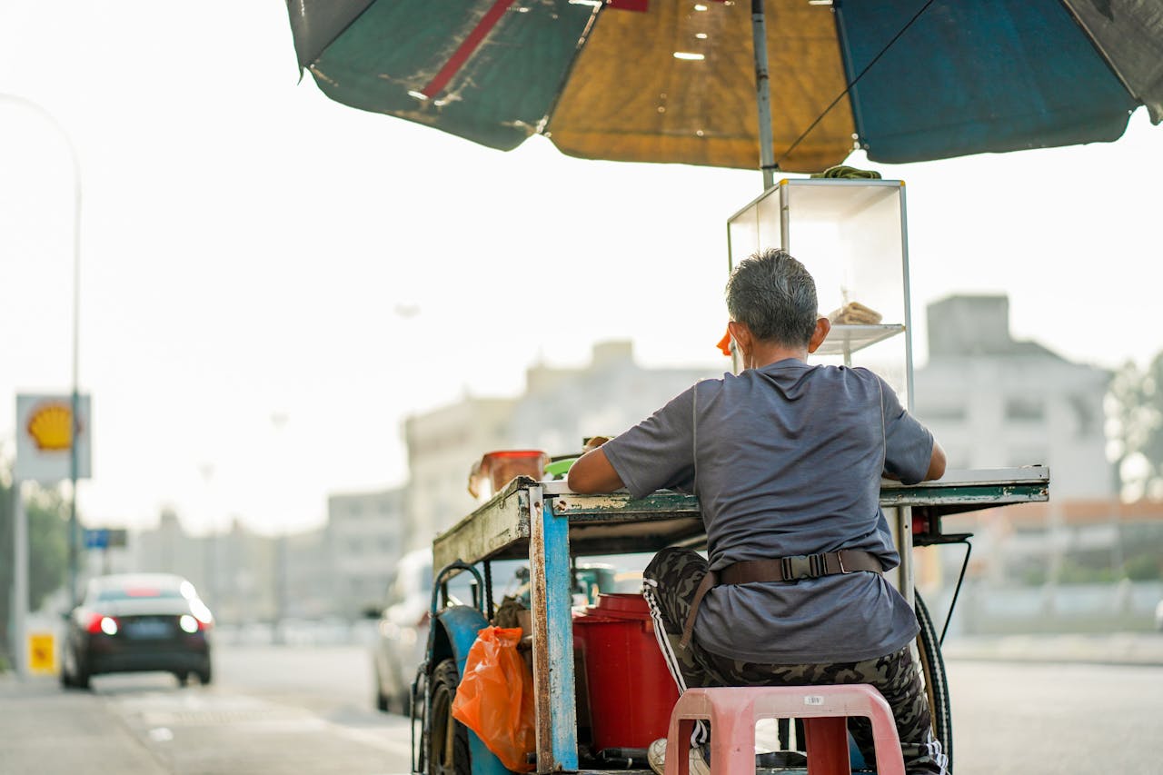 Elderly vendor under an umbrella on city streets of Klang, Malaysia. Captured from behind.