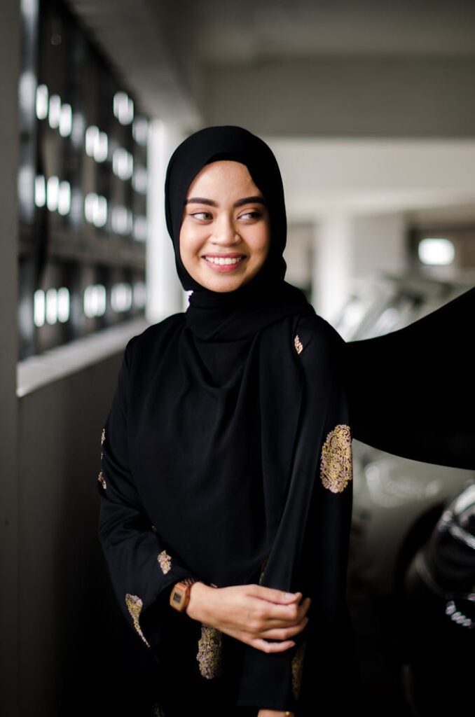 A woman in a black hijab smiles warmly while standing indoors, showcasing elegance and confidence.